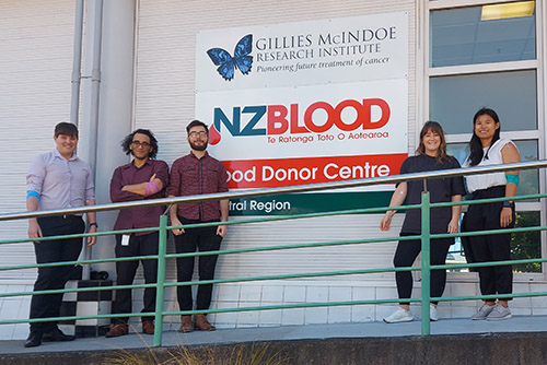 Five smiling coworkers with bandages on their arms stand in front of the New Zealand Blood Donor Centre